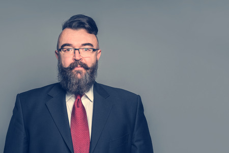 Adult bearded man in a suit, glasses and a mohawk hairstyle on a gray backgroundの写真素材