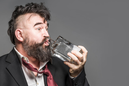 Disheveled bearded man in suit drinks alcohol from a glass bottle on a gray backgroundの写真素材
