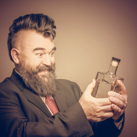 Smiling adult bearded man in suit looking at a bottle of alcohol in his hand on a brown backgroundの写真素材