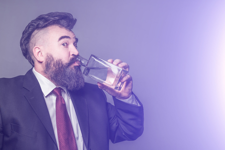 Adult bearded man in suit drinking alcohol from a glass bottle on a purple backgroundの写真素材