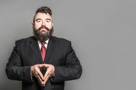 Adult bearded man in a suit with folded triangle hands on a gray backgroundの写真素材