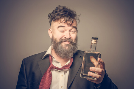 Disheveled smiling bearded man in suit holding in his hand a bottle of alcohol. Tonedの写真素材