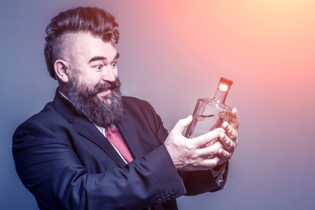 Happy adult bearded man in suit looking at a bottle of alcohol in the sunlightの写真素材