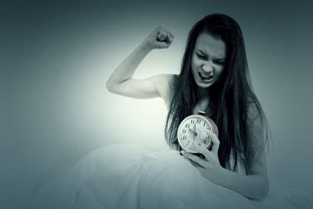 The woman is annoyed, trying to bring down the alarm. good morning. The brunette holds an alarm clock in her hands, sitting on the bed. I wake up from the alarm clock. Not getting enough sleep.の写真素材
