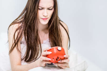 The brunette holds an alarm clock in her hands, sitting on the bed. Set an alarm before bed.の写真素材