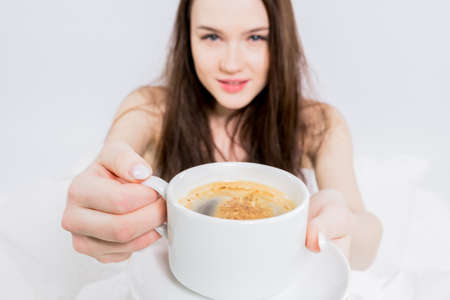 Tasty breakfast. White bedroom. good morning. The brunette holds a cup of hot drink, offers to try....