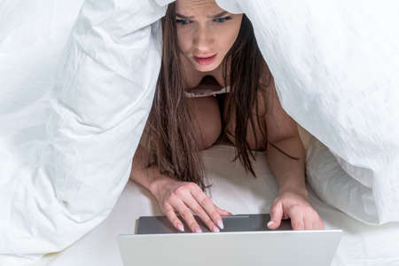 The brunette is covered with a blanket over her head and looks in a frightened way at the laptop....