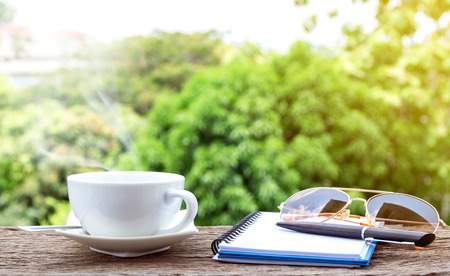Hot coffee cup  with a notebook, sunglasses and pen on the old wooden floor with colorful green nature background.の写真素材