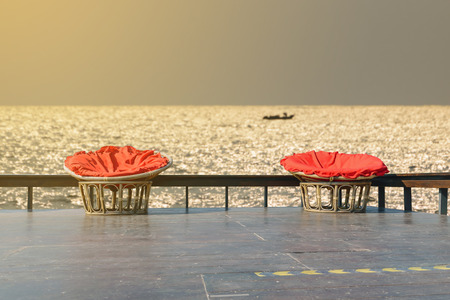 Chairs made of rattan and upholstery fabrics in red on wooden terrace with the backdrop of the ocean.の写真素材