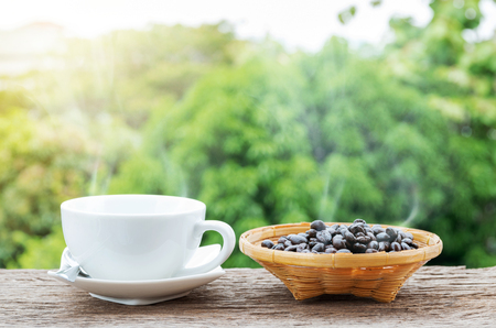 Hot coffee cup and coffee beans in the bamboo plate on the old wooden floor with colorful green nature background.の写真素材