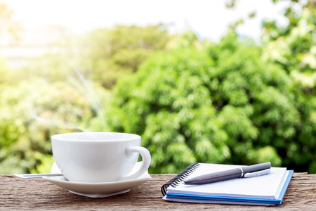 Hot coffee cup  with a notebook and a pen on the old wooden floor with colorful green nature background.の写真素材