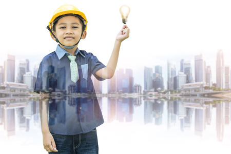 Double exposure of the Asian boy dressed as an engineer wearing a safety hat holding a light bulb on a cityscape backdrop-Imagination and creativity concepts.の写真素材