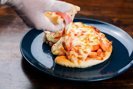 Close-up of hand wearing rubber gloves picking up pizza slice in a ceramic plate on a wooden table.の写真素材