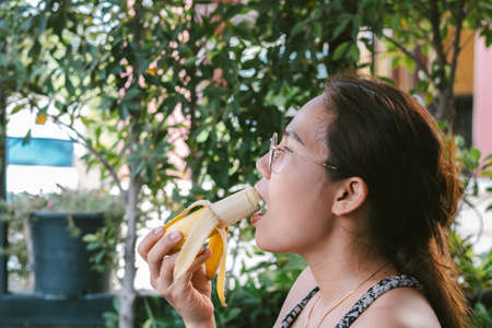 Close-up of a face of an Asian woman in glasses eating a ripe bananaの写真素材