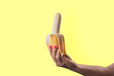 Closeup a woman's hand painted red nails holding a ripe banana isolated on yellow pastel colors backgroundの写真素材