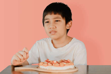 Asian cute boy sitting at the dining table with pizza on the table in front on pink color background.の写真素材