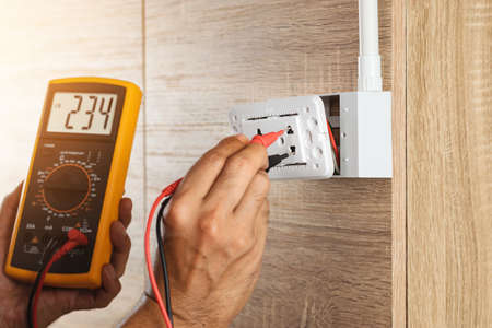 Electrician using a digital meter to measure the voltage at a wall socket on a wooden wall.の写真素材