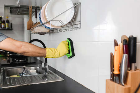 Close up human hand in a yellow rubber glove is using a plastic brush to scrub the tile wall in the kitchen room.の写真素材