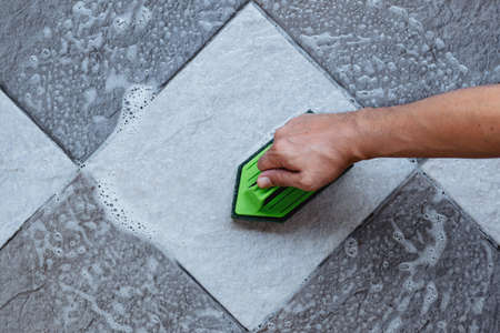 Top view of a human hand are using a green color plastic floor scrubber to scrub the tile floor with...