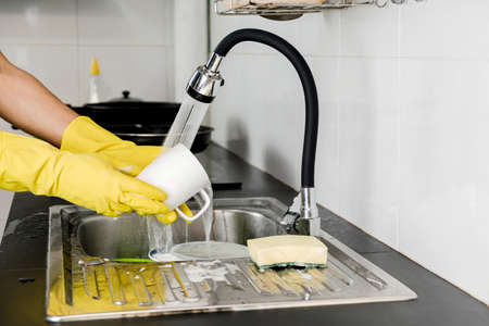 Close up human hands in yellow rubber gloves washing a ceramic coffee mug in the kitchen sink.の写真素材
