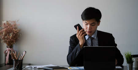 Businessman in suit talking on mobile phone and working on tablet computer at office desk.