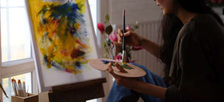 A young woman artist holding paintbrush and painting with oil paint on canvas at her workshop. Hobby...