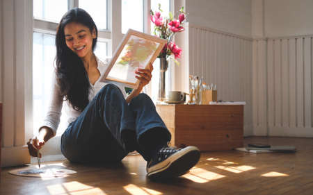Happy female Artist holding framed canvas and paintbrush ready for painting at home during weekend.の写真素材