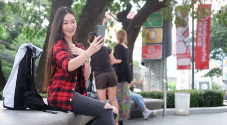Young Asian woman in street style clothes taking selfie with smart phone while sitting at outdoor.の写真素材