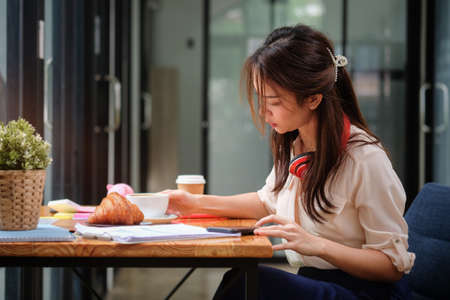 Side view female office worker using smart phone and reading business documents at office desk.の写真素材