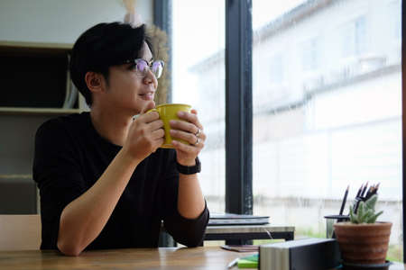Thoughtful young asian man holding cup of coffee and looking out of window.