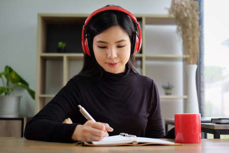 Young asian woman listening to music with wireless headphone and writing some information on...