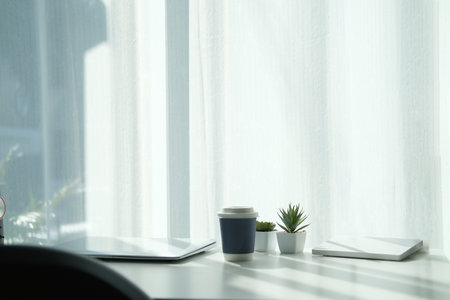 Comfortable workplace with laptop computer, cup of coffee and houseplant on white table near window.の写真素材