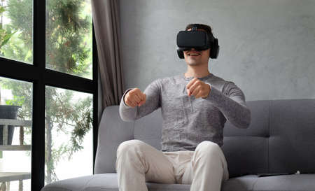 Young man in casual clothes sitting on sofa and playing video games with virtual reality headset.