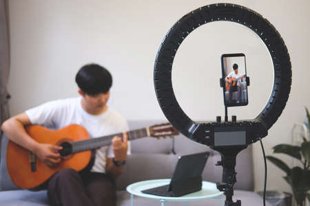 Young asian man playing acoustic guitar and recording video on smartphone.