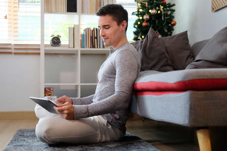 Handsome man in casual clothes sitting in living room and surfing internet with digital tablet.の写真素材