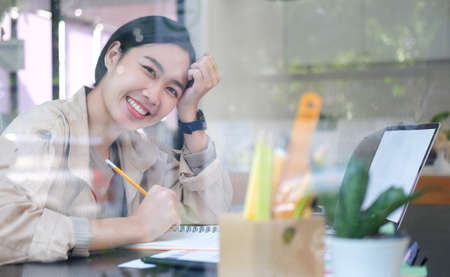 View through glass window of cheerful asian woman sitting in creative office and smiling to camera.の写真素材