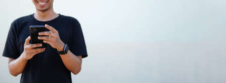 Smiling young asian man using smart phone while standing against white wall.