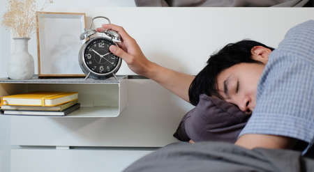 Sleepy man lying on bed and turning off alarm clock switch in the morning.の写真素材