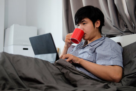 Smiling handsome man drinking coffee and using laptop computer on bed.の写真素材