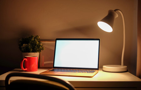 Computer laptop with blank screen, coffee cup and lamp on white table in dark room.の写真素材