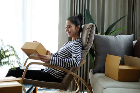 Happy young woman sitting in living room and opening parcel box. Online shopping and delivery...
