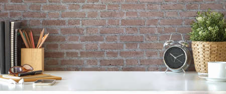 Stylish workplace, alarm clock, houseplant, books and pencil holder on white table with brick wall.