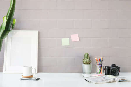Stylish workplace with empty picture frame, cactus, camera and coffee cup on white table.