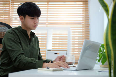 Young asian man holding coffee cup and working with laptop computer at home office.の写真素材