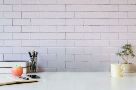 Books, smart phone, apple, coffee cup and potted plant on white table with brick wall.