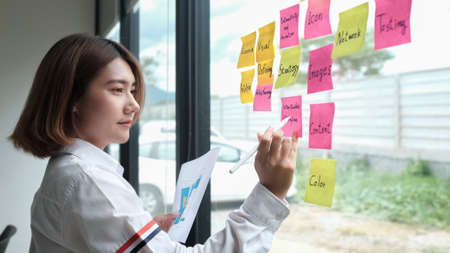 Focused young businesswoman writing new ideas on sticky notes on glass wall.