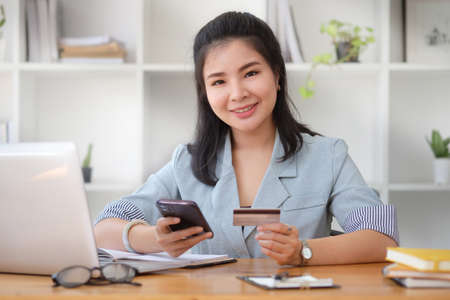 Cheerful young asian woman holding credit card and smart phone for shopping online.