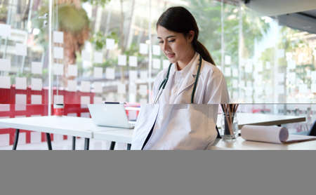 Smiling female doctor in white uniform using digital tablet while standing at medical office....