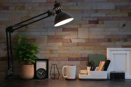 Comfortable workplace with stationery, coffee cup, lamp and picture frame on black table with brick wall.の写真素材