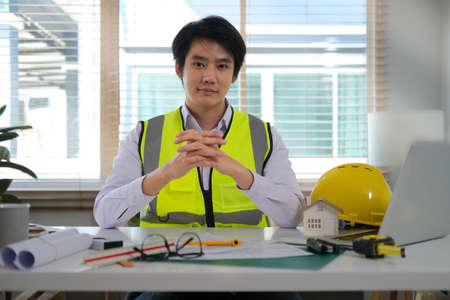 Young Asian Engineer in formal suit and hardhat with rolled blueprints sitting at workplace and smiling to camera.の写真素材